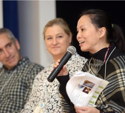 A woman on the right speaks into a microphone, holding a booklet. Two people seated beside her listen attentively. All are in casual attire with lanyards, suggesting a vibrant panel discussion or engaging conference session atmosphere.