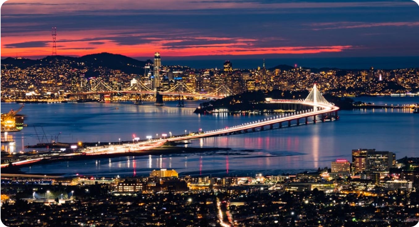 A panoramic view of a lit-up cityscape at dusk, featuring a brightly illuminated bridge that facilitates the evening transit, spanning the shimmering water. The skyline, dotted with glowing buildings, is set against a vibrant sunset with shades of red and orange.