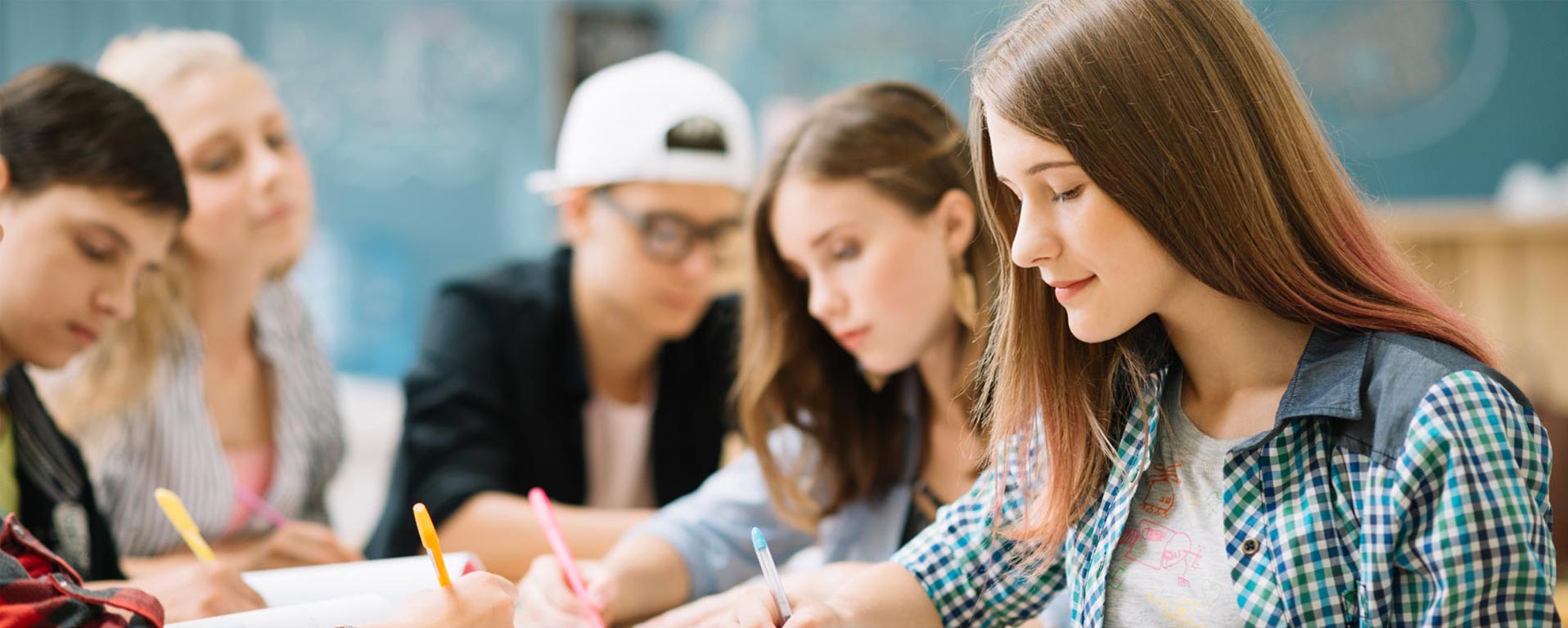 A group of students sits at a desk in a classroom, intently focused on writing and studying with notebooks and pens in hand. Surrounded by essential resources, like textbooks and the chalkboard in the background, they immerse themselves in learning.