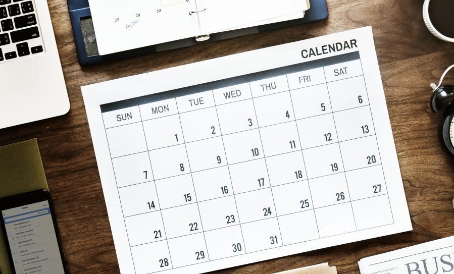 A monthly calendar lies on a wooden desk alongside a laptop, notebook, and headphones. The calendar's grid, styled like shuttle routes, is labeled Sunday to Saturday with numbers from 1 to 30, illustrating the month’s journey.