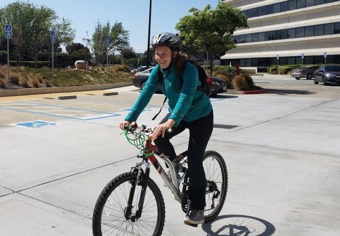 A person wearing a helmet and teal jacket is joyfully riding a mountain bike in a parking lot. Among the backdrop of cars and a large building, this cheerful commuter embraces the freedom of two wheels.