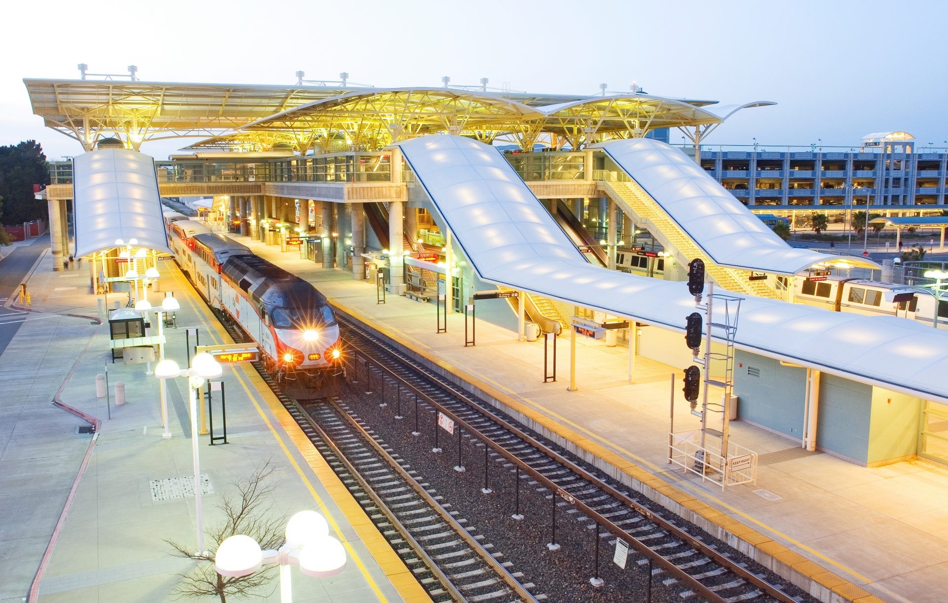 Millbrae Station shines at dusk, featuring a modern design with several covered platforms and a parked train. Bright lights illuminate the scene, highlighting the multi-level parking garage in the background.