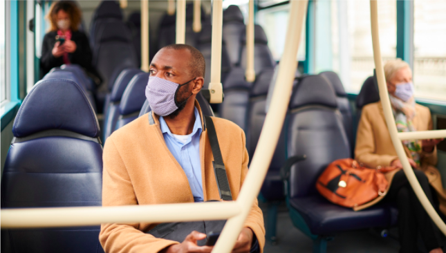 A man wearing a light purple face mask and camel coat sits on a bus, reflecting on the changes in commuting after COVID. In the background, two other masked passengers are seated. The bus appears mostly empty, with blue seats.