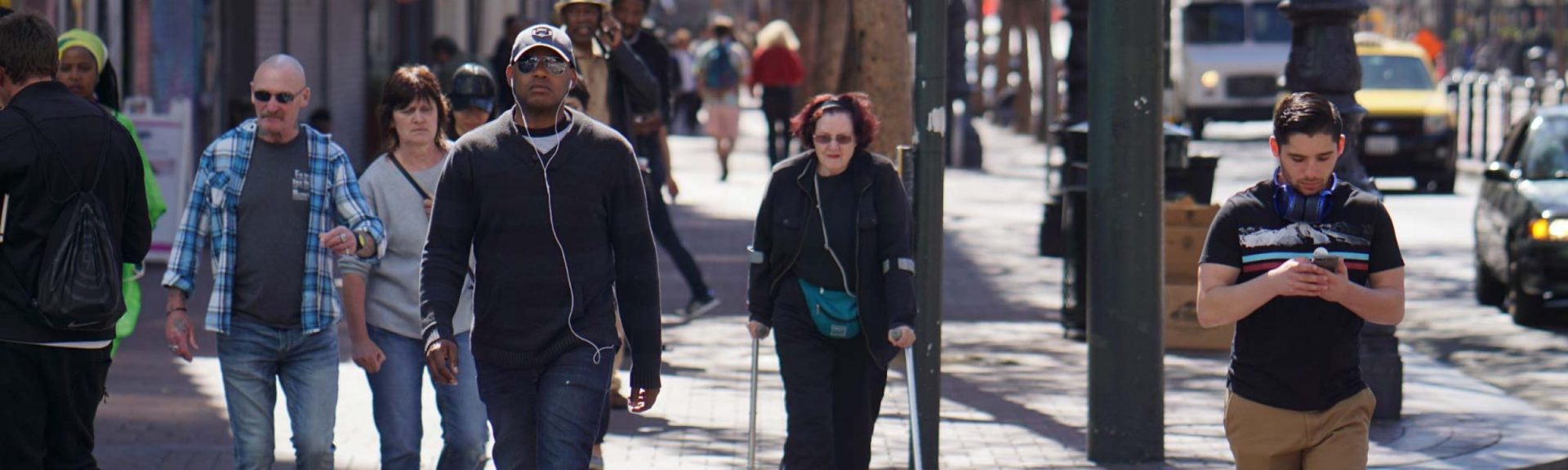 A diverse group of commuters walks along a city street. Some are dressed casually, others more formally. One person uses a cane, while another checks their smartphone. Trees and buildings form the backdrop.