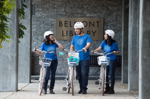 Three people in blue shirts and white helmets stand with bicycles in front of the "Belmont Library." They are smiling, engaged in conversation. Through TDM Grants, they support eco-friendly travel solutions. Each bike sports a basket, symbolizing community efforts for a sustainable future.