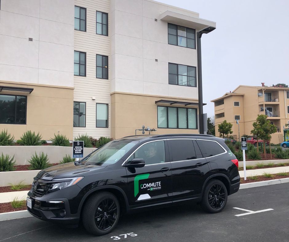 A black SUV with a "Commute" logo is parked in front of a modern apartment building, symbolizing accessibility for workforce housing. The vehicle rests in a designated space, with lush greenery and residential structures forming the backdrop.