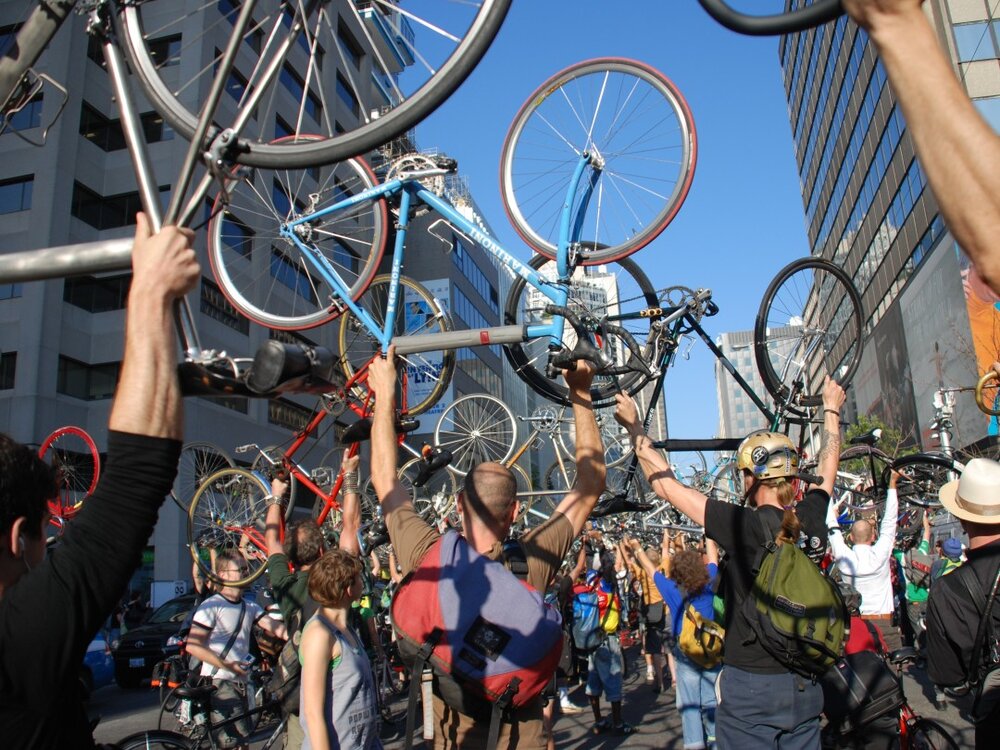 Amidst a backdrop of modern buildings and a clear blue sky, a vibrant crowd gathers on the city street, hoisting bicycles aloft in celebration. This lively scene underscores the city's commitment to initiatives like workforce housing and TDM grants, bringing the community together.
