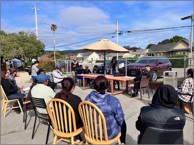 A group of people are seated in a semicircle in an outdoor setting, engaging in a discussion. Some are on chairs, others on benches surrounding a table with papers detailing outreach and next steps. An umbrella provides shade to the table. Houses and power lines are visible in the background.