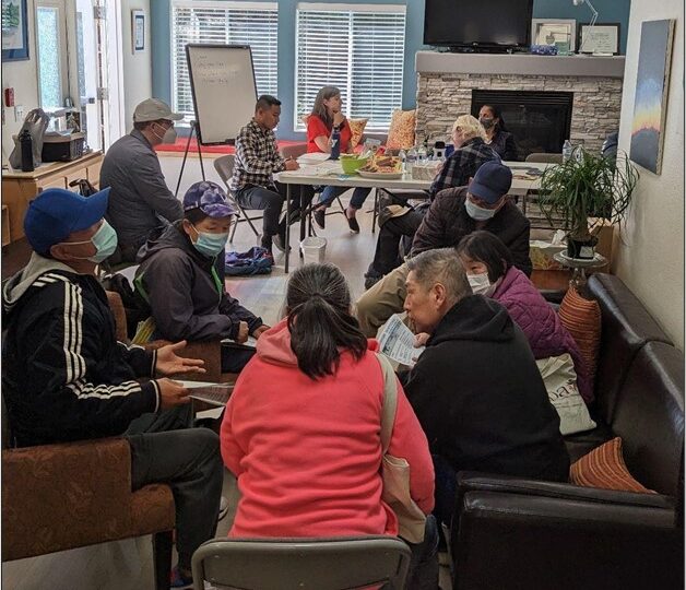 A group of people are seated in a room, engaged in conversation about equity outreach. Most are wearing masks. Some are sitting on couches, while others occupy chairs around a table. A whiteboard and a fireplace are visible in the background as sunlight streams through the windows.