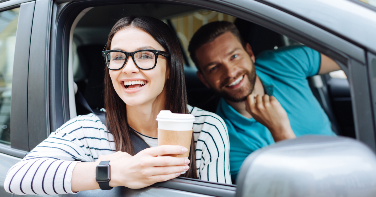 A woman wearing glasses and a striped shirt is smiling while holding a coffee cup out of a car window. A man in a blue shirt sits beside her, also smiling, with his hand on his neck. Both appear happy and relaxed inside the parked car.
