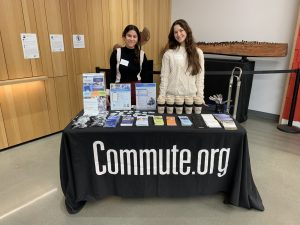 Two people stand behind a table with a black "Commute.org" tablecloth, offering insights into TDM strategies. The table displays brochures, flyers, reusable cups, and pens in a modern indoor setting with a light wood-paneled wall as the perfect recap backdrop.