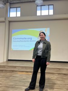 A person stands smiling in front of a presentation screen displaying a slide titled "Commute.org Overview/Descripción General" during a Community Workshop. The room, promoting Climate Resilient Communities, has high ceilings and wooden floors.