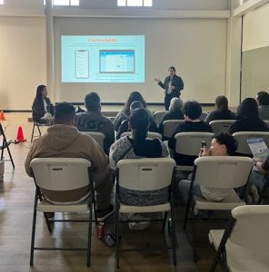 Audience seated in a hall attentively listening to a presenter. The presenter stands beside a large screen displaying a "Commutestar" slide with mobile app visuals during the Community Workshop. The room boasts high ceilings and an abundance of natural light from large windows.