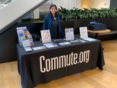 A person stands behind a table covered with TDM resources for employers—brochures, flyers, and buttons. The table has a black cloth with "Commute.org" printed on it. The setting appears to be indoors, near some plants and a staircase.