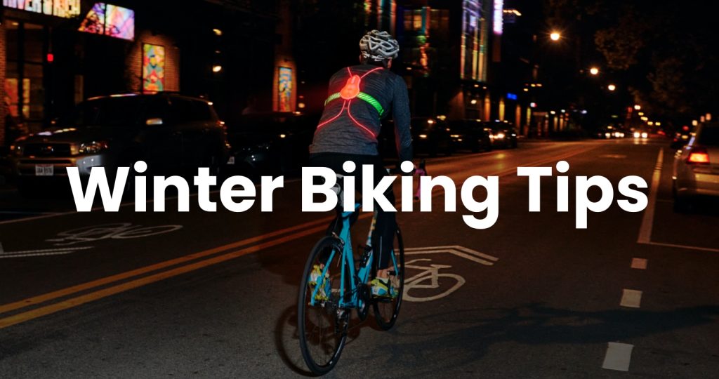 A cyclist wearing a helmet and a jacket with LED lights rides at night on a city street in San Mateo County. Text reads "Winter Biking Tips" across the image, helping commuters stay safe this season.