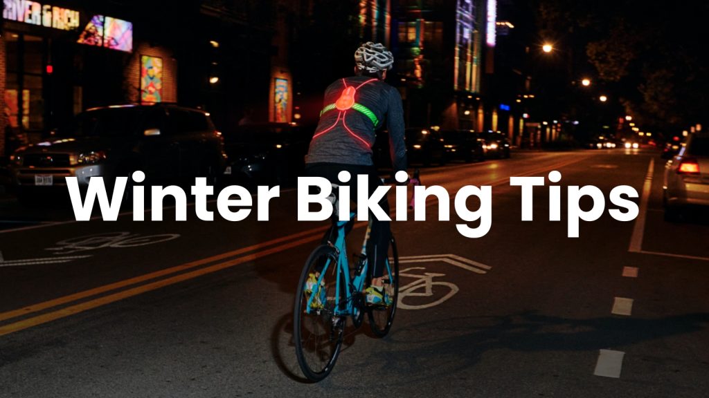 A cyclist wearing a helmet and a jacket with LED lights rides at night on a city street in San Mateo County. Text reads "Winter Biking Tips" across the image, helping commuters stay safe this season.