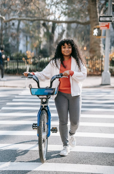 A young woman with curly hair, dressed in a white jacket and red shirt, walks a blue bicycle across a crosswalk in a city park area on a sunny day, enjoying the rewards for commuters who choose cycling.