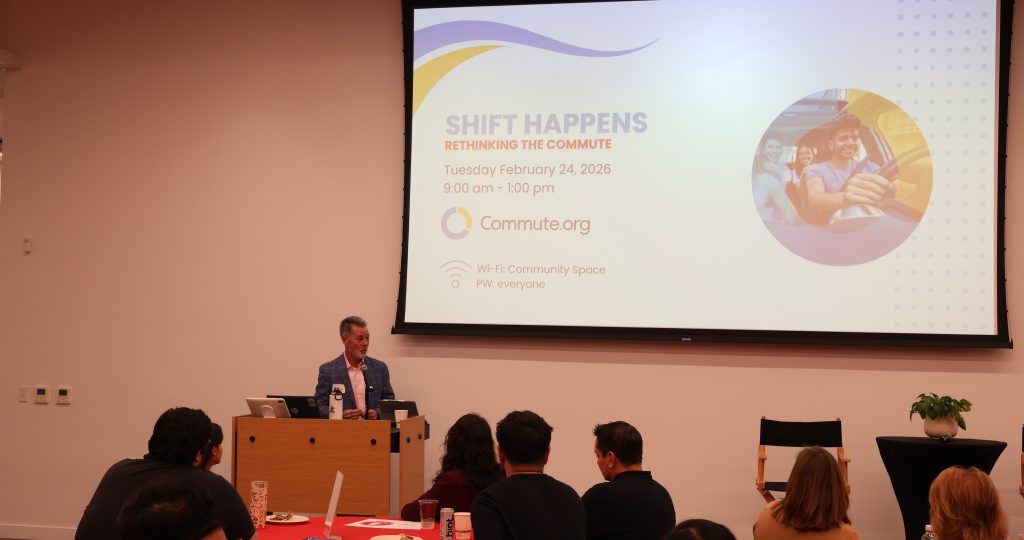 A speaker stands at a podium in front of an audience, presenting a slideshow titled "Shift Happens: Rethinking Commute," with event details and a photo of people in a car displayed on a large screen behind him.