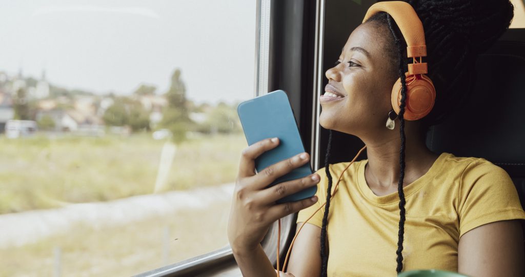 A woman wearing orange headphones and a yellow shirt smiles while looking out the window of a moving train, holding a blue smartphone—perhaps checking the 2025 Commuter Challenge Results Are In! A green travel mug sits in the foreground.